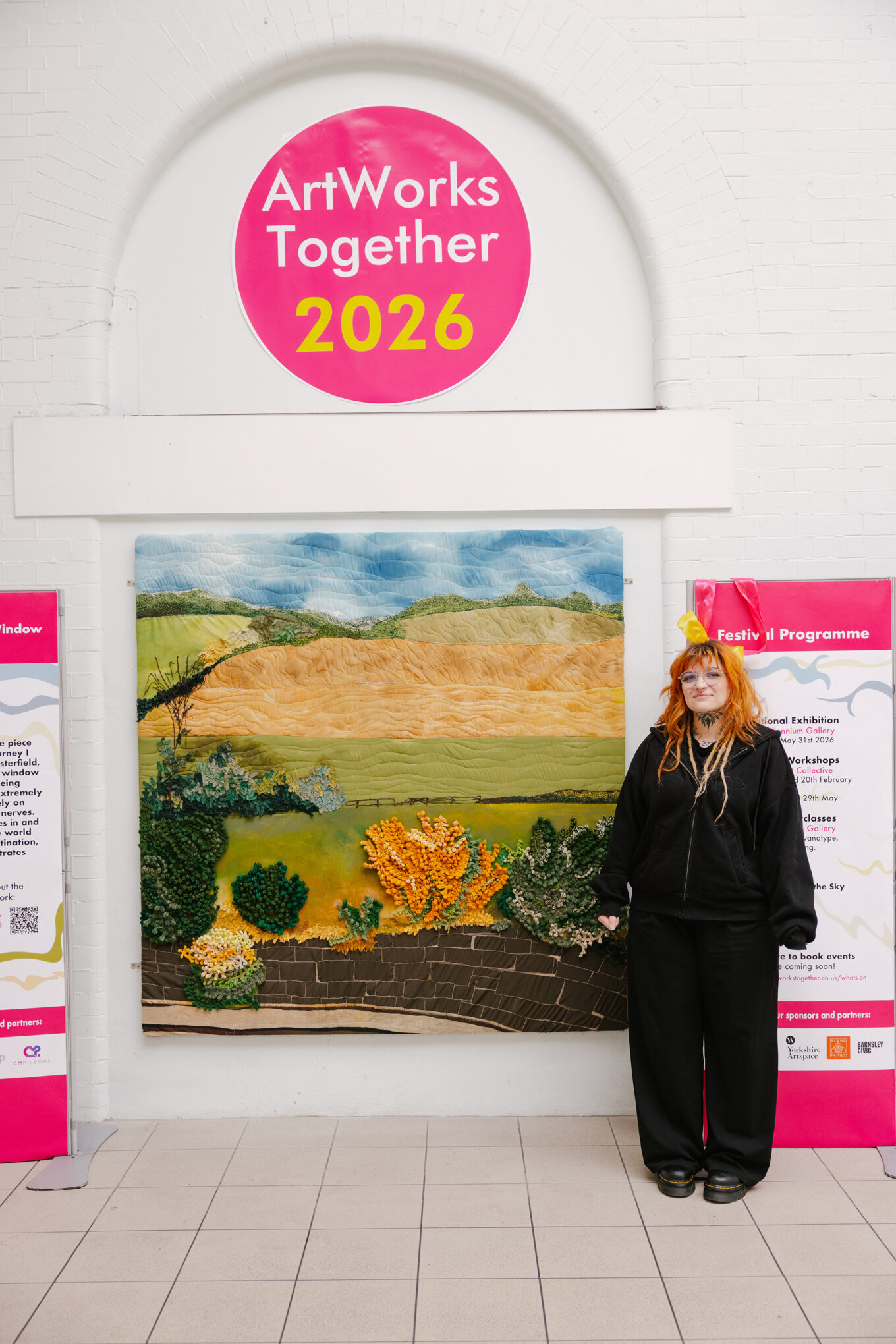 A woman wearing black with long dyed orange hair is standing next to her artwork, shown in the lobby of a train station. It shows a view of the country from a train window, with fields, bushes and a train platform in the foreground.
